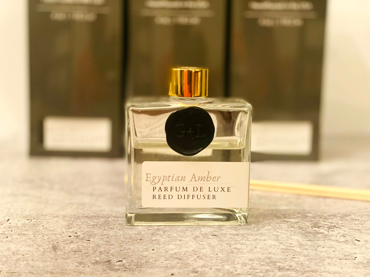 Clear reed diffuser with gold cap on a wooden surface, with blurred boxes in the background.