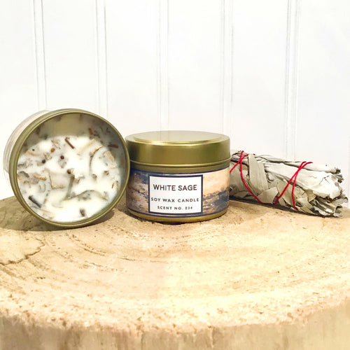 White Sage soy wax candle in a gold tin with a white label, surrounded by white sage smudge sticks on a wooden surface.
