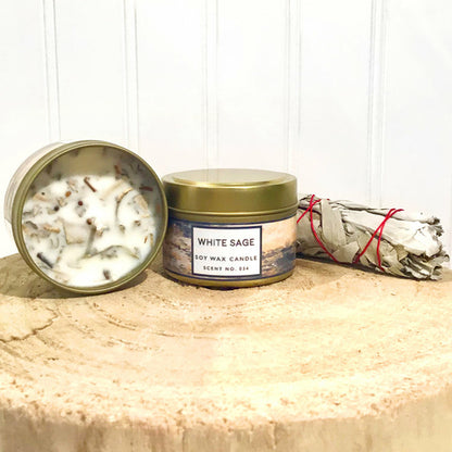 White Sage soy wax candle in a gold tin with a white label, surrounded by white sage smudge sticks on a wooden surface.
