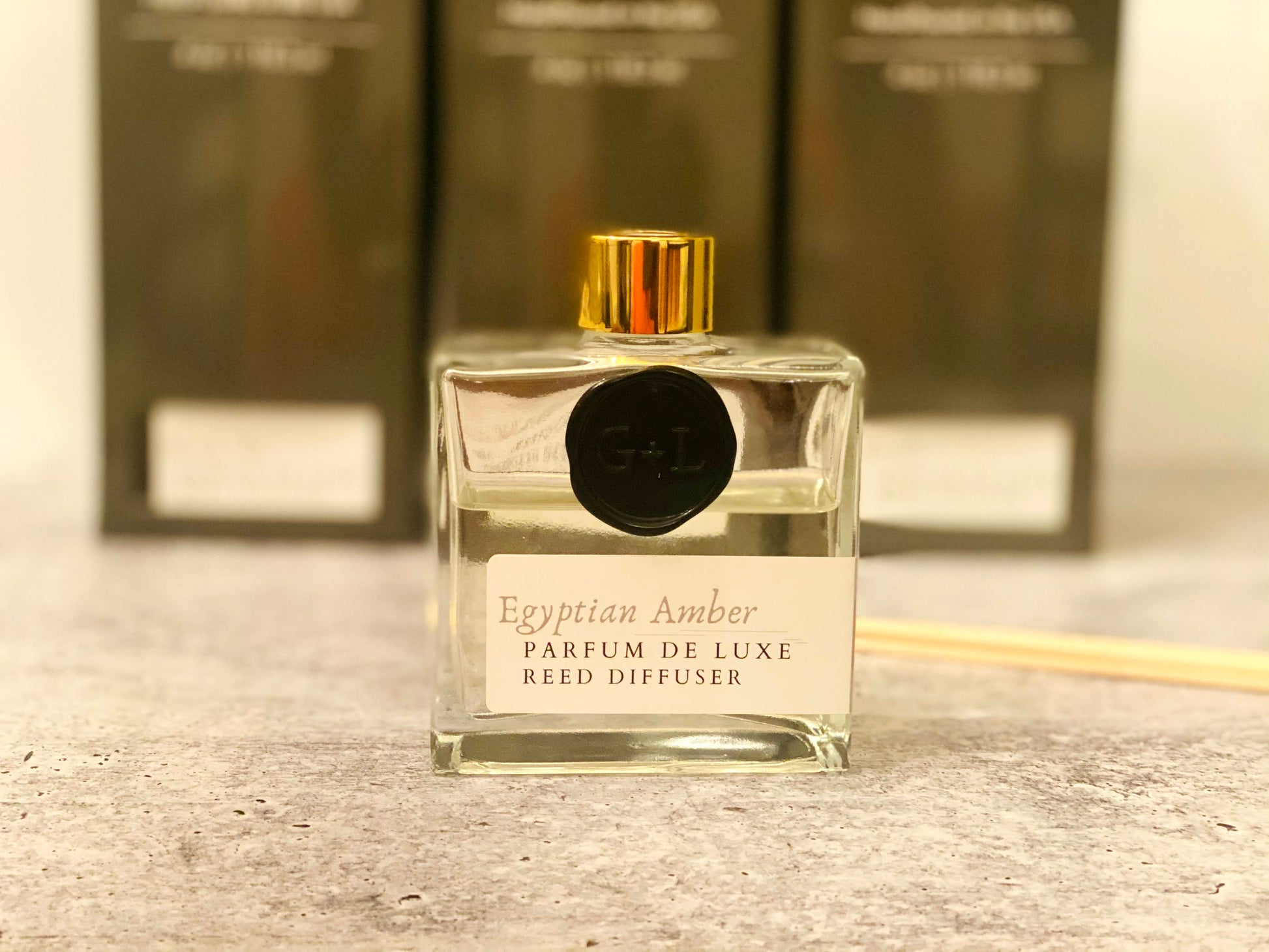 Clear reed diffuser with gold cap on a wooden surface, with blurred boxes in the background.