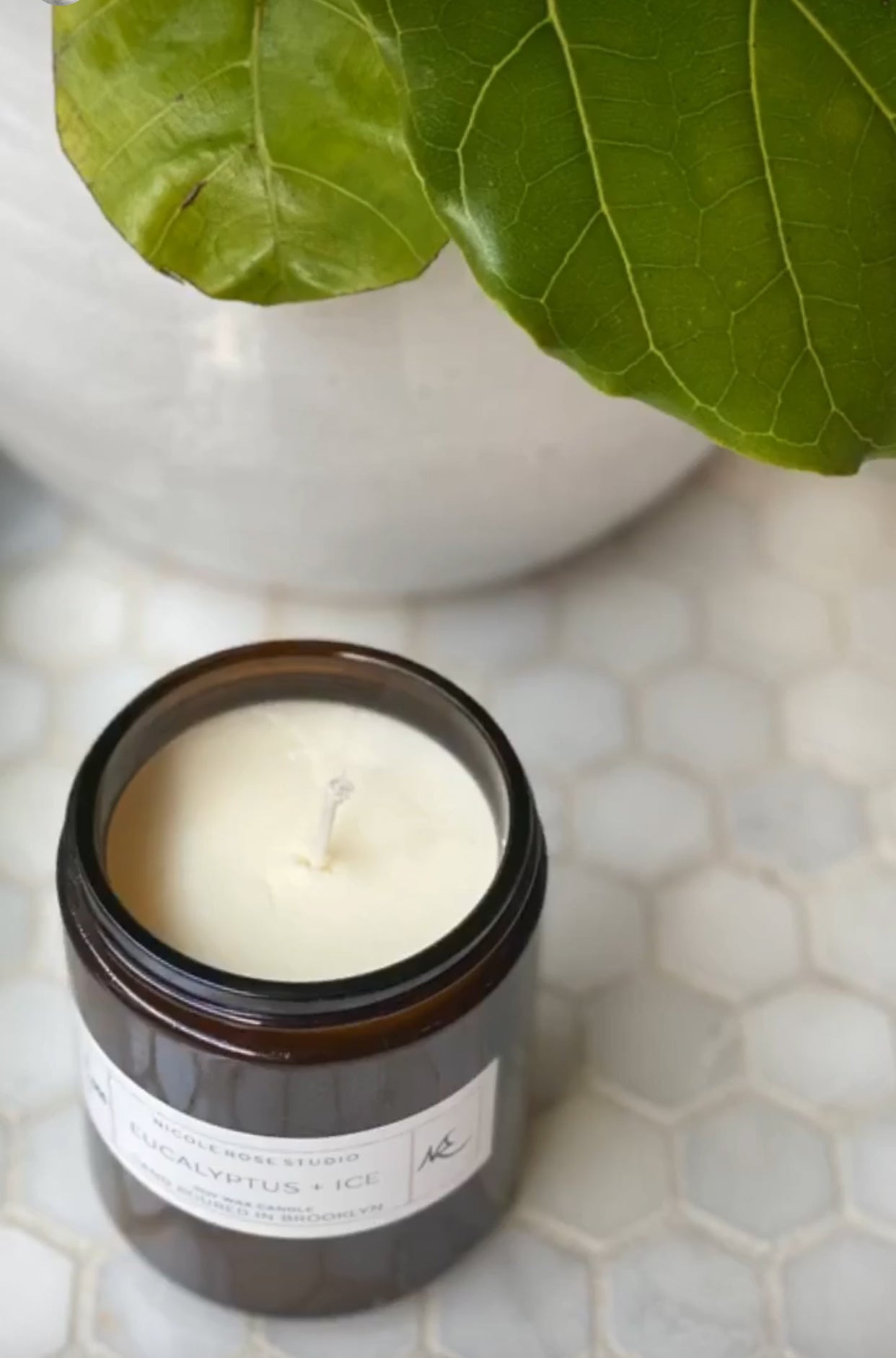 Candle in a glass jar on a tiled surface with green leaves in the background