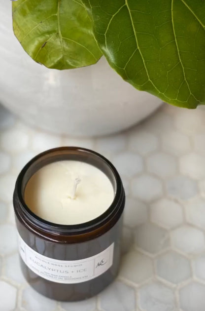 Candle in a glass jar on a tiled surface with green leaves in the background