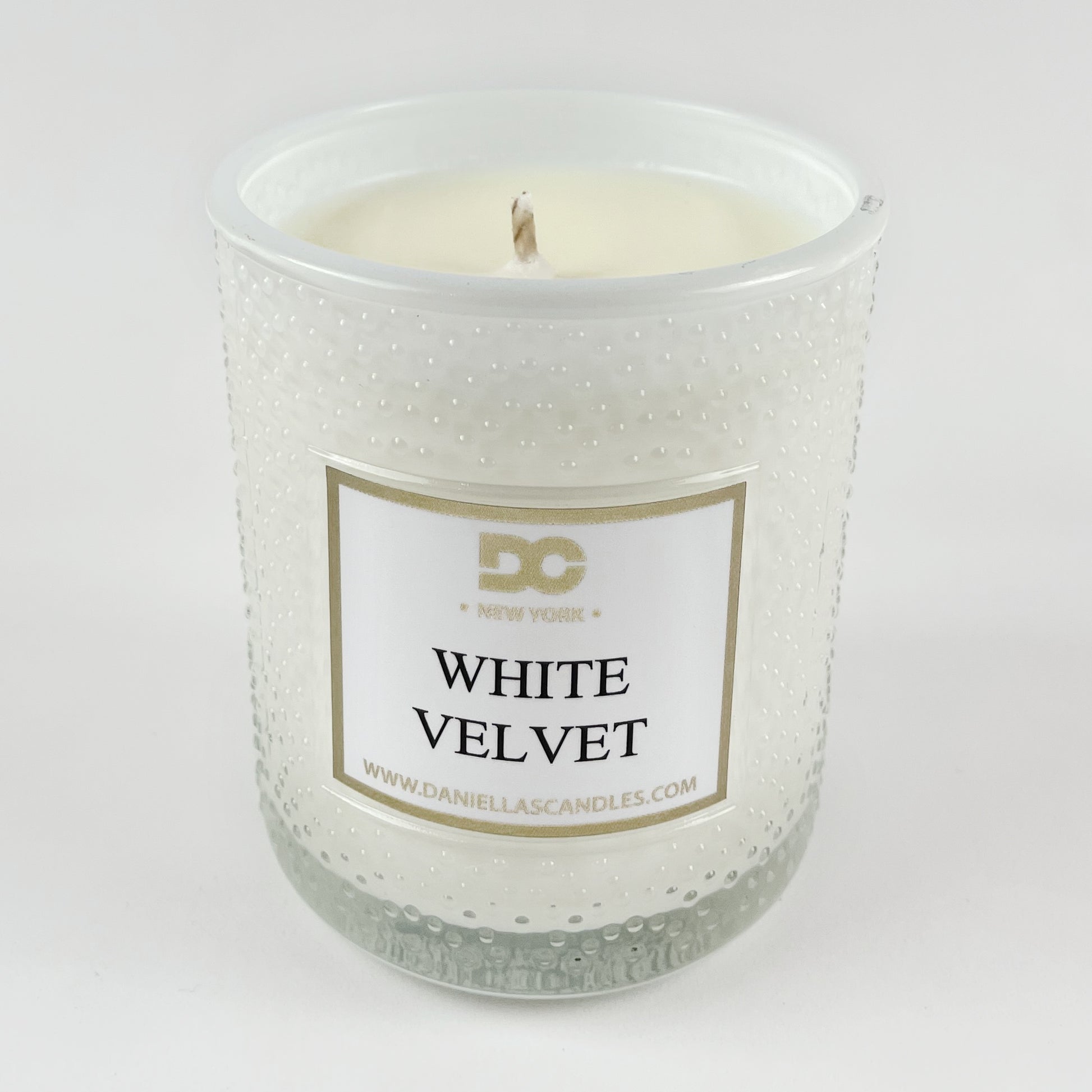 White candle in a textured glass jar with a label on a white background