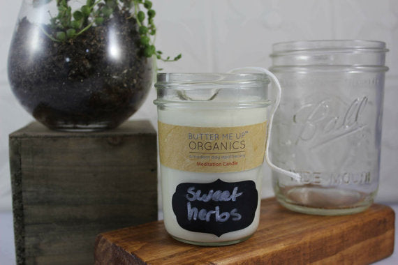 Butter Me Organics candle labeled 'Sweet Herbs' on a wooden surface with a glass jar and plant in the background.