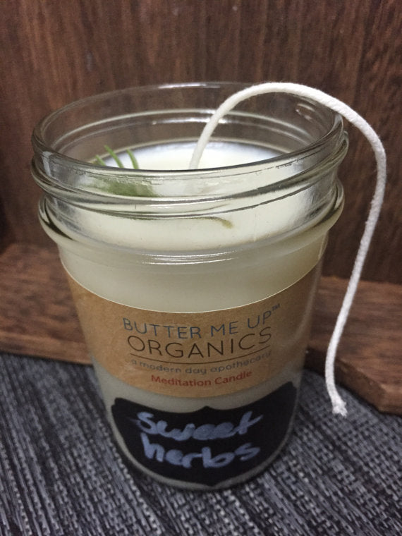Candle in a glass jar labeled 'Butter Me Up Organics Sweet Herbs' on a wooden surface.