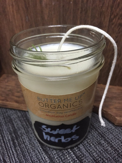 Candle in a glass jar labeled 'Butter Me Up Organics Sweet Herbs' on a wooden surface.