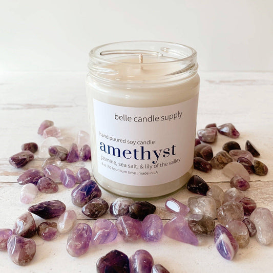 Candle labeled 'amethyst' from belle candle supply surrounded by amethyst stones on a light wooden surface.