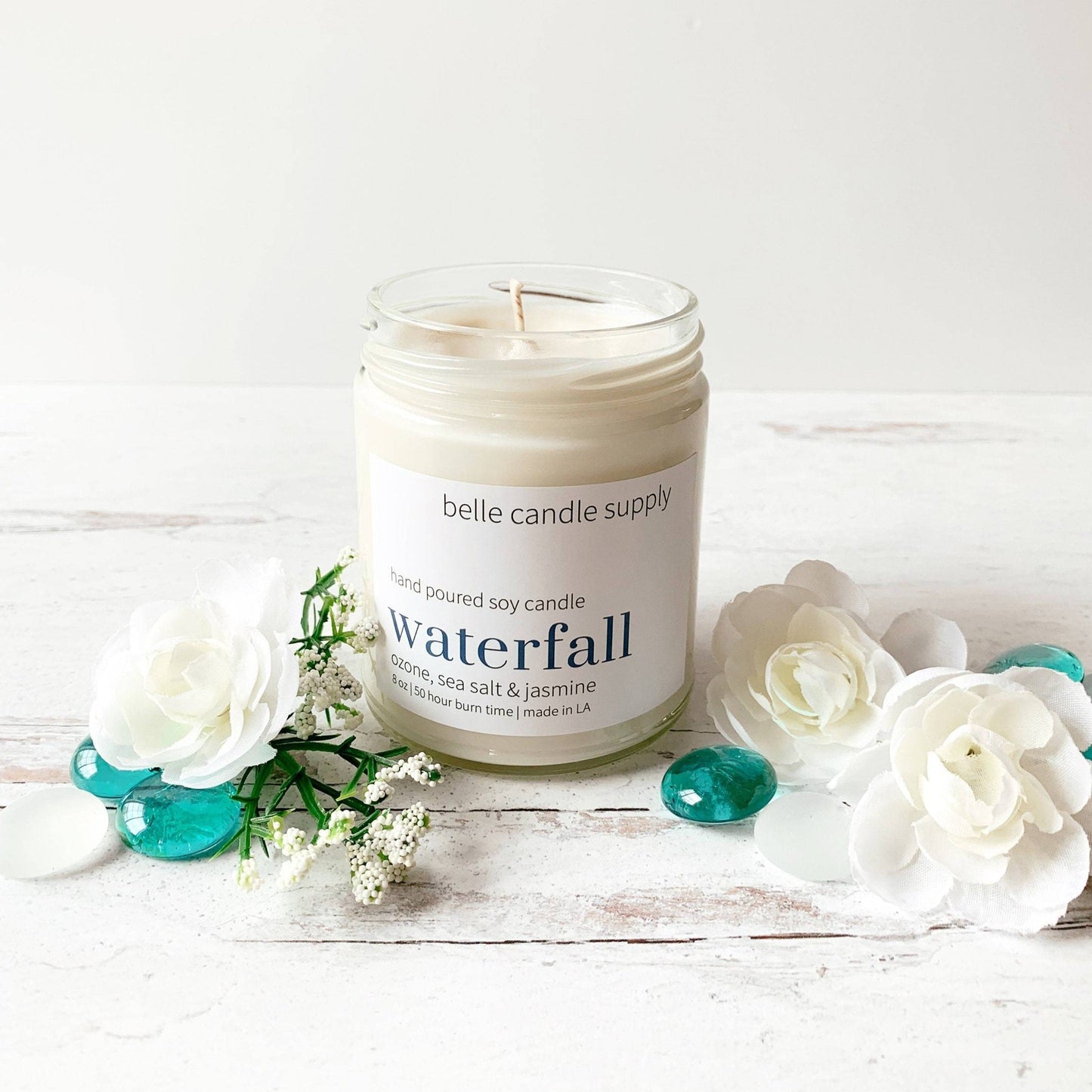 Candle labeled 'Waterfall' from belle & candle supply on a wooden surface with flowers.