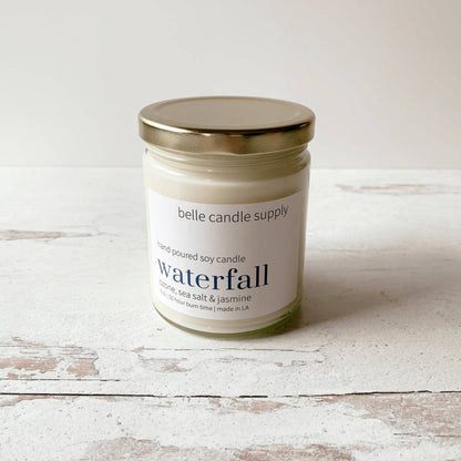 Candle jar labeled 'belle candle supply' with 'waterfall' fragrance on a wooden surface.