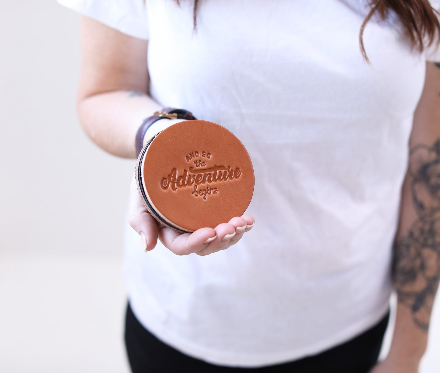 Person holding a round brown leather item with 'Live Life Adventure' text on a plain background